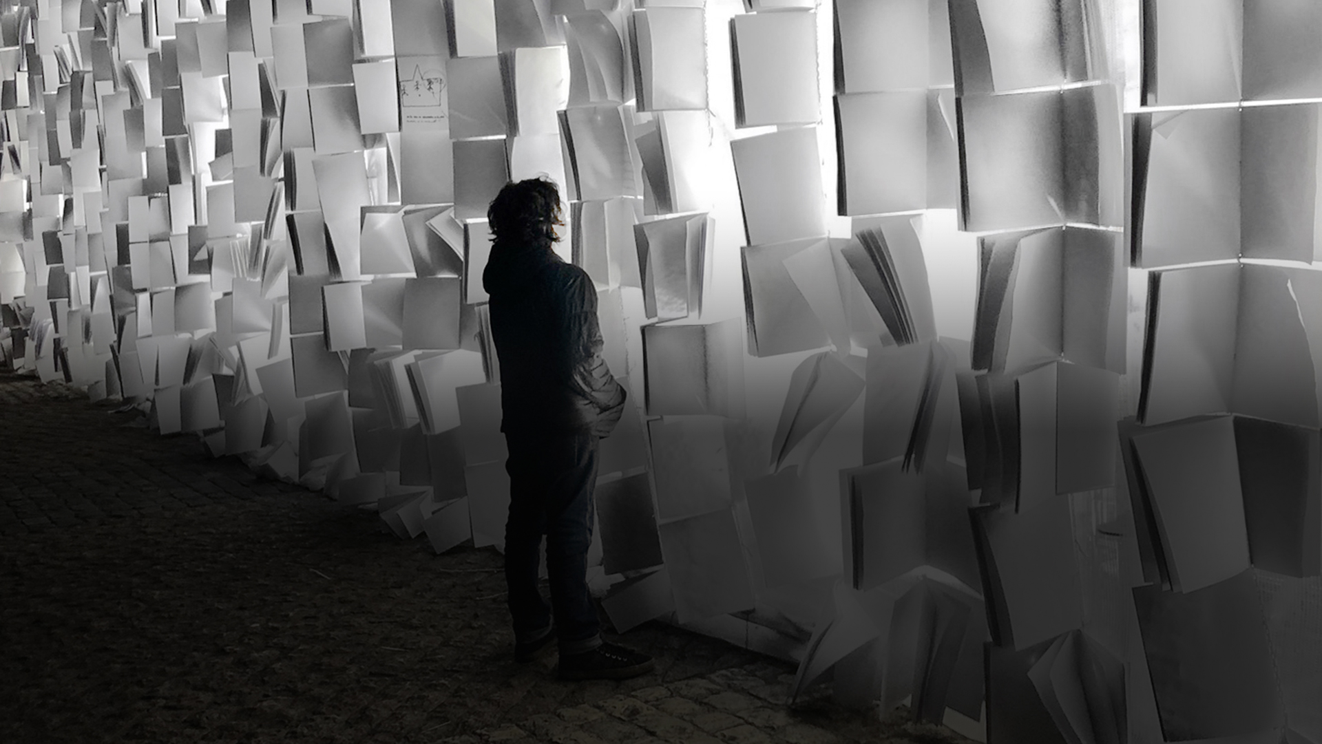 a young person standing in front of a lighted wall made of blank page notebooks